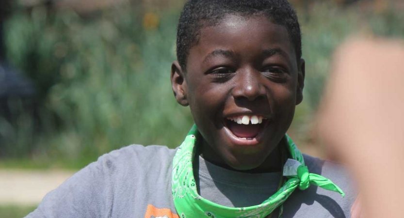 A young person smiles a big smile. There is blurry greenery in the background. 